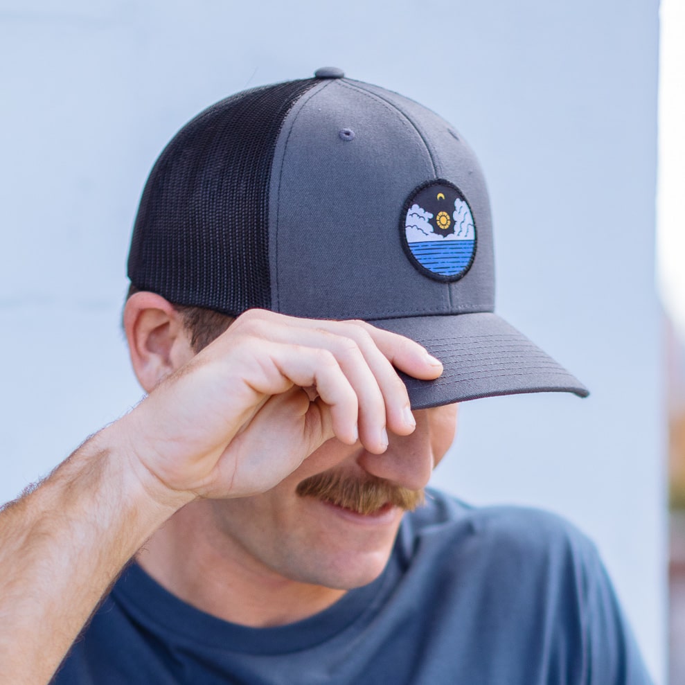 Man wears a grey trucker hat with a patch sewn on it